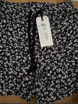 Svarta blommiga shorts från Lindex stl xs - Säljer ett par svarta shorts från Lindex med vitt blommigt mönster och snörning i midjan. De är tillverkade i 100% viskos och har en skön loose passform. Perfekta för varma dagar och har en mjuk känsla mot huden.