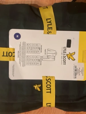 Mörkgrön rutig Pyjamas Sätt från Lyle & Scott - Snygg mörkgrön Pyjamas Sätt från Lyle & Scott med rutigt mönster och klassisk gul logga på bröstet. Västen har knappar framtill och en bröstficka. Perfekt för dig som gillar stilrena detaljer och vill sticka ut lite extra.   (HELT OANVÄNT OCH OÖPPNADE) Ny pris 1000Kr