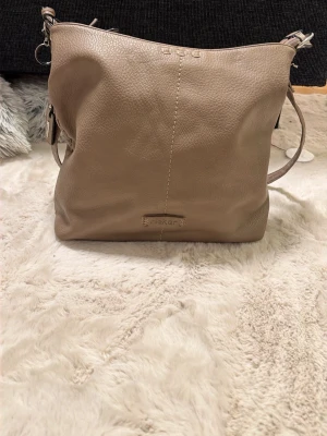 Beige axelväska från Rieker i skinn - Säljer en beige axelväska från Rieker i mjukt skinn med justerbar rem. Väskan har en enkel design med en dragkedja på baksidan och ett ljust, blommigt foder med logotyp. Perfekt storlek för vardagsprylar och har snygga sömdetaljer framtill.