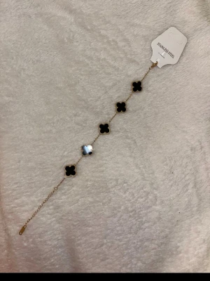 Svart blommigt armband i stål - Snyggt armband med fem svarta blommor i fyrklöverform, infattade i guldfärgad kedja. Armbandet är tillverkat i rostfritt stål och har en tunn, elegant kedja med justerbar längd. Perfekt för att ge din outfit en cool och stilren touch.