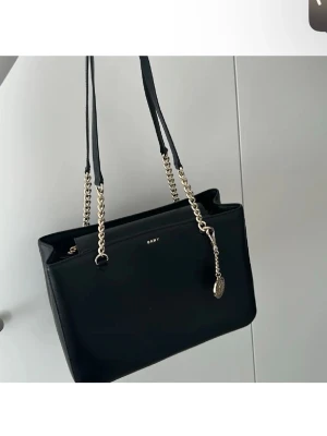 Svart axelväska från DKNY med kedja - Snygg svart axelväska från DKNY med stilrena guldfärgade kedjor som axelremmar. Väskan har en klassisk rektangulär form och är tillverkad i slätt skinn. Diskret DKNY-logga framtill och dekorativt hänge i metall. Perfekt för dig som gillar en clean och modern look.