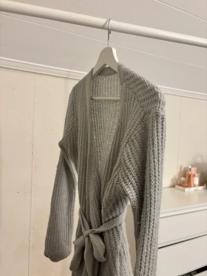 Grå stickad kofta med knytband - Mysig grå stickad kofta med lång ärm och knytband i midjan. Koftan har en öppen front och grovstickad struktur som ger en avslappnad vibe. Perfekt att slänga över en t-shirt eller linne när det blir lite kyligt.