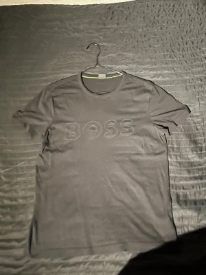 Svart BOSS t-shirt med tryck - Svart t-shirt från BOSS med diskret BOSS-tryck på bröstet och liten logga i nacken. Klassisk rund hals och korta ärmar. Materialet är mjuk bomull som känns skön mot huden. Perfekt för dig som gillar stilrena och enkla plagg med en touch av premium. Storlek S med en regular pass form