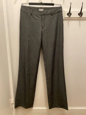 Grå randiga kostymbyxor Madlady - Snygga grå kostymbyxor från Madlady i extra lång modell, storlek 36. Byxorna har smala vita ränder och klassisk rak passform med hällor för bälte. Perfekt för dig som gillar stilrena och trendiga plagg med lite edge. Midjan är midwaist/low waist och innebens längden är ca 90 cm men går att sy upp om det behövs.