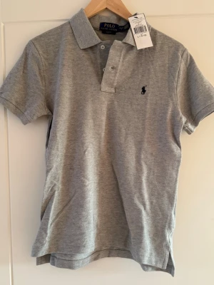 Grå piké t-shirt från Polo Ralph Lauren - Säljer en stilren grå piké t-shirt från Polo Ralph Lauren i slim fit. T-shirten har korta ärmar, klassisk krage och knappar framtill. Materialet är mjuk bomull och den har den ikoniska Polo-loggan broderad på bröstet. Perfekt för en clean och chill look.