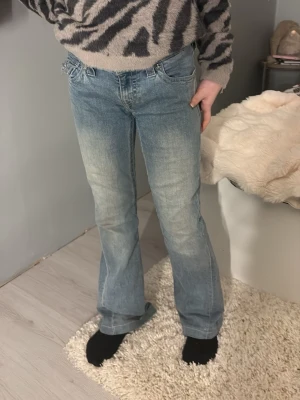 True Religion ljusblå bootcut jeans - Säljer ett par ljusblå jeans från True Religion med klassiska broderade fickor och silverfärgade knappar. Modellen har bootcut-snitt och normal passform, tillverkade i jeansmaterial. Snygga detaljer på bakfickorna och ikonisk patch i midjan.