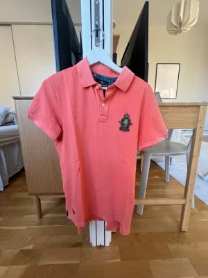 Korallfärgad piké från Morris - Säljer en snygg korallfärgad pikétröja från Morris Gent's med klassisk krage och korta ärmar. Pikén har broderad logga på bröstet och ett stort broderat emblem på ryggen. Materialet är bomull och insidan av kragen har ett mönstrat tyg i blått och orange. använd vid några tillfälle men i väldigt fint skick. 