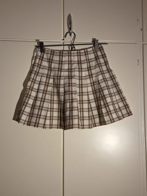 Brun och vit rutig kortkjol från H&M - Säljer en brun och vit rutig kortkjol från H&M Divided i storlek 34. Kjolen har plisserad design och är gjord av 98% polyester och 2% elastan, vilket gör den mjuk och bekväm. Perfekt för dig som gillar preppy stil och vill ha något snyggt till garderoben.