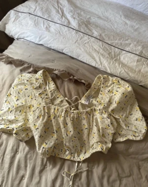 Blommig blus med snörning från Zara - Säljer en söt blommig blus från Zara i storlek M. Blusen är vit med gula och svarta blommor och prickar, har puffiga korta ärmar och snörning framtill. Croppad modell med elastisk kant nedtill och vid halsen. Perfekt för sommarens alla tillfällen!