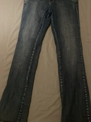 Blå bootcut jeans  - Säljer ett par blå jeans med bootcut jeans med coola paljettdetaljer på fickorna. Jeansen har en mörk tvätt med slitningar och klassiska sömmar. Säljer då de inte passar längre. De står size 5 men skulle säga att de passar någon som bär xs/s. Som ni ser på bilderna är jeansen lite slitna men inget man tänker på när man har på sig de men därav priset💕 skriv vid eventuella frågor eller liknande 