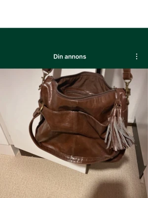 Brun axelväska med tofsar i skinn - Säljer en stor och mjuk brun axelväska i skinn med flera fack och dragkedja upptill. Väskan har en bred axelrem och dekorativa tofsar i skinn på sidan. Perfekt för dig som vill ha plats för allt och gillar en klassisk look.