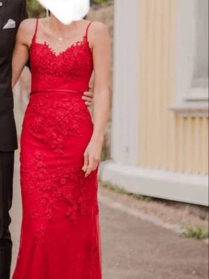 Röd balklänning  - Säljer en elegant röd långklänning med smala axelband och v-ringad urringning. Klänningen har vackra blommiga spetsdetaljer och ett smalt bälte i midjan. Perfekt för bal eller fest!