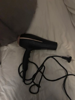 Svart hårfön med roségulddetaljer - Snygg hårfön i matt svart med stilrena roségulddetaljer längs sidorna och runt luftintaget. Ergonomiskt handtag och lång sladd med EU-kontakt. Utrustad med flera värme- och hastighetsinställningar. Perfekt för att fixa frisyren snabbt hemma.