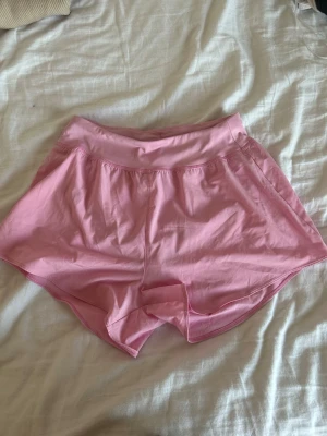 Rosa träningsshorts från SOC - Säljer ett par ljusrosa träningsshorts från SOC i storlek 38. De har en loose passform, elastisk midja och är gjorda i ett lätt syntetmaterial med meshfoder för extra ventilation. Perfekta för löpning eller gymmet under varma dagar.