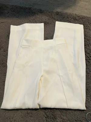 Vita kostymbyxor med hög midja - Snygga vita kostymbyxor med hög midja och raka ben. Byxorna har stilrena pressveck framtill och är gjorda i ett slätt material som ger en clean look. Perfekta för dig som gillar minimalistisk stil och vill ha ett par klassiska byxor i garderoben.