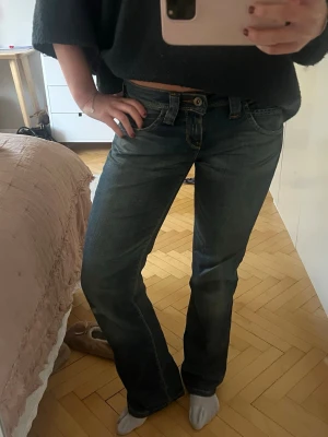 Blå bootcut jeans från Lee - Säljer ett par klassiska blå jeans från Lee med bootcut-snitt och låg midja. Jeansen har snygga kontrastsömmar, fem fickor och Lee-logga på bakfickan. Materialet är slitstarkt jeans och passformen är normal. Perfekt för dig som gillar retrovibe och bekväma jeans.