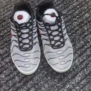 Nike Air Max Plus Tn grå/svarta sneakers - Nike Air Max Plus Tn sneakers i grått och svart med röda detaljer. Skorna har vågformade linjer på ovandelen, svart snörning och en tydlig Tn-logga bak. Ovandelen är i syntetmaterial och mesh för en sportig vibe. Perfekta för dig som gillar streetwear och snygga detaljer.