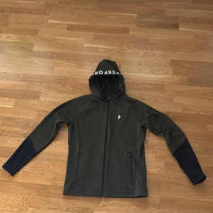Grön hoodie från Peak Performance S - Mörkgrön hoodie från Peak Performance med hel dragkedja, huva med vit text och två fickor med dragkedja. Insidan är fodrad med mjuk fleece och ärmarna har svarta detaljer. Perfekt för kyliga dagar och outdoor-aktiviteter.