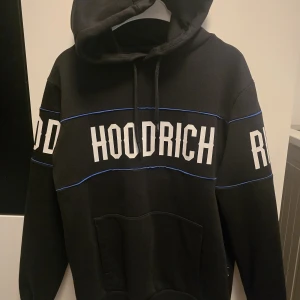 Svart HOODRICH hoodie med blå detaljer - Svart hoodie från HOODRICH i storlek XS med stora vita tryck på bröst, rygg och ärmar. Snygga blå linjer som kontrast och klassisk känguruficka framtill. Huva med snörning och mjukt material som passar perfekt till streetwear-stilen.