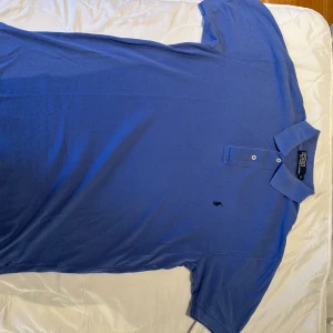 Polo Ralph Lauren - Klassisk blå pikétröja från Polo Ralph Lauren med broderad logga på bröstet. Tröjan har korta ärmar, krage och två knappar framtill. Tillverkad i mjuk bomull som känns skön mot huden. Perfekt för en clean och stilren look. Hör av dig vid fler frågor 