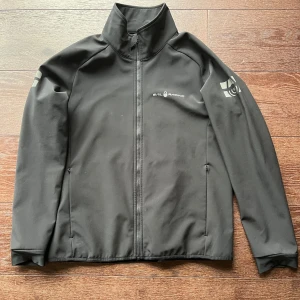 Svart vindjacka från Sail Racing - Svart vindjacka från Sail Racing med hög krage och hel dragkedja framtill. Jackan har diskreta loggor på bröstet, ärmen och ryggen. Jackan har strl 170 men är som en XS. Sonen som haft den var 170-170 cm när han kunde ha den.