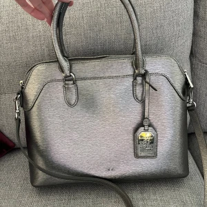 Silverfärgad handväska från Ralph Lauren - Snygg handväska i metallic silver från Ralph Lauren. Väskan har både handtag och axelrem, dragkedja upptill och ett rymligt foder med logotypmönster. Äkta läder och har en metallbricka med RL-logga framtill. Köptes på Zalando för några år sedan men bara stått i garderoben, kanske använd max 5 gånger men är i nyskick 😊