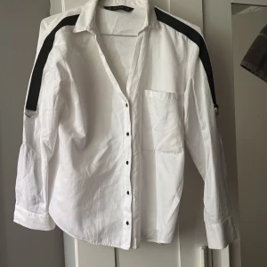 Vit skjorta med svarta detaljer Zara - Säljer en vit skjorta från Zara Basic i bomull med svarta band längs axlarna och cool metallring på ena ärmen. Skjortan har knappar framtill, bröstficka och lång ärm. Snygg oversized passform som ger en chill vibe.
