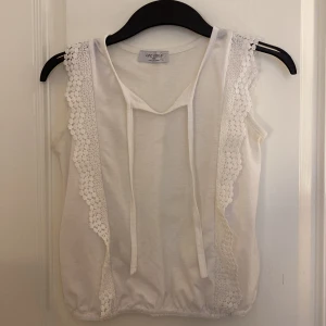 Vit spetsdetaljerad linnetopp LPC Girls - Supersöt vit linnetopp från LPC Girls med breda spetsdetaljer längs axlar och sidor. Toppen har knytband framtill och är ärmlös med lätt rynkad nederkant. Perfekt för en fräsch och somrig look.