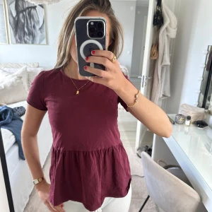 Vinröd peplum blus i bomull - Säljer en vinröd peplum blus med korta ärmar och rund halsringning. Toppen har en volang nedtill som ger en snygg och avslappnad siluett. Tillverkad i mjuk bomull, perfekt för dig som gillar en enkel men trendig look.