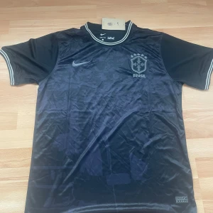 Svart Brasilien fotbollströja Nike XL - Snygg svart Brasilien fotbollströja från Nike i Dri-FIT-material. Tröjan har korta ärmar, diskret camo-mönster på framsidan, silverfärgade detaljer och broderat CBF-märke. Ribbstickade muddar och krage med vita ränder ger en clean look. Slim fit size gör tröjan som en L-M i storlek