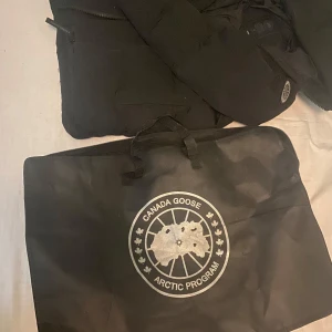 Svart dunjacka från Canada Goose - Säljer en svart dunjacka från Canada Goose, modell macmillan S/P. Jackan har huva, dragkedja och  Canada Goose-logga på ärmen. Perfekt för kalla dagar och riktigt snygg streetstil. Kommer med  fodral.