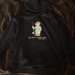Svart hoodie Polo Bear Ralph Lauren - Svart hoodie från Polo Ralph Lauren med ikoniska Polo Bear-trycket framtill. Tröjan har huva, stor magficka. Har aldrig använt den och köpte den ny, tuvär så har jag inte lappen men har påsen den kommer i