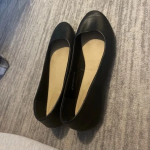 Svarta ballerinaskor i skinnimitation - Klassiska svarta ballerinaskor med rund tå och platt sula. Skorna har en enkel och stilren design, perfekta för dig som gillar minimalistisk stil. Utsidan är i skinnimitation och insidan är ljusbeige för extra kontrast.