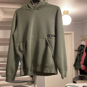Grön hoodie från Sail Racing - Snygg grön hoodie från Sail Racing med huva och stor magficka. Tröjan har en stilren look med diskret logga på bröstet och är perfekt för dig som gillar en avslappnad stil. Mjuk insida och ribbade muddar vid ärmslut och nederkant. Nästan aldrig använd