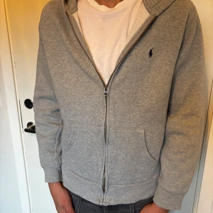 Grå hoodie från Polo Ralph Lauren XL - Snygg grå hoodie från Polo Ralph Lauren i storlek XL(Barnstorlek) Tröjan har huva, dragkedja framtill och två stora fickor. Klassisk logga broderad på bröstet. Mjuk insida i bomullsmix, perfekt för chill dagar. Passar dig som gillar enkel och stilren streetwear.