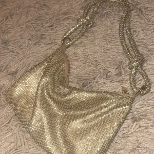 Glittrig väska  - Superfin axelväska i guld med glittrande stenar över hela väskan och ett coolt axelband med knutdetaljer. Perfekt för att sticka ut och ge din outfit extra bling. Väskan stängs med dragkedja och har en mjuk, flexibel form.
