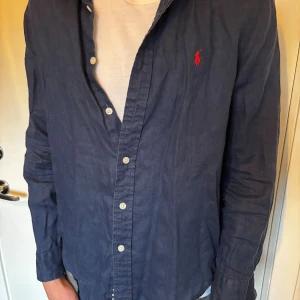 Mörkblå skjorta från Polo Ralph Lauren - Säljer en mörkblå skjorta från Polo Ralph Lauren med klassisk krage och knappar framtill. Skjortan har lång ärm och den ikoniska röda loggan broderad på bröstet. Tillverkad i bomull, perfekt för en clean och stilren look.