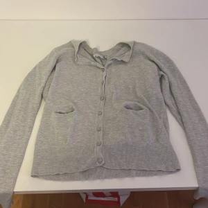 Grå kofta från 157 med knappar - Ljusgrå kofta från 157 i mjuk bomullsmix. Klassisk modell med långa ärmar, ribbade muddar och knäppning framtill. Två fickor framtill ger en chill vibe. Perfekt att slänga över en t-shirt eller linne.