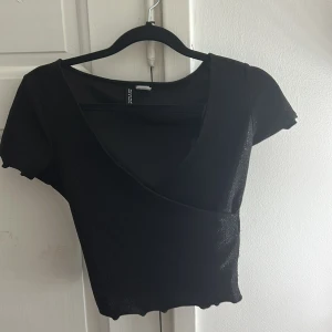 Svart glittrig omlottopp Divided XXS - Snygg svart croppad omlottopp från Divided i storlek XXS. Toppen har korta ärmar, glittrigt tyg och en v-ringad omlottdesign som ger en trendig look. 