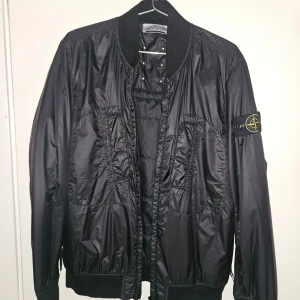 Svart bomberjacka från Stone Island - Säljer en svart bomberjacka från Stone Island med klassisk patch på vänster ärm. Jackan har ribbade muddar, dragkedja framtill och är tillverkad i glansig polyester. Snygg och stilren modell som passar perfekt till streetwear. Flera fickor framtill och coola detaljer vid axlarna. Priset kan diskuteras vid snabb affär 