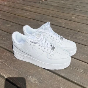 Nike Air Force 1 helvita sneakers - Klassiska Nike Air Force 1 sneakers i helvitt skinn med perforerad tå, snörning och låg profil. Snygg och tidlös modell med rund tå och platt sula, perfekt till streetwear eller casual outfits. Diskret Nike-logga på sidan och bak.