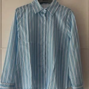 Ny. Blåvit randig skjorta med knappar - Snygg blå skjorta med vita vertikala ränder och klassisk krage. Skjortan har långa ärmar, knappar framtill och vid ärmslut. Perfekt för en fräsch och stilren look. Materialet känns som bomull och är lätt och bekvämt.