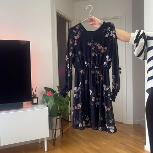 Mörkblå blommig klänning med resår - Supersöt mörkblå klänning med blommigt mönster i rosa och beige toner. Klänningen har lång ärm med resår vid midjan och ärmsluten, vilket ger en snygg siluett. Perfekt för dig som gillar romantiska vibes och vill ha något bekvämt men ändå stilrent.