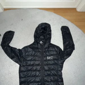 Svart jacka från EA7 Emporio Armani - Svart pufferjacka från EA7 Emporio Armani med huva och dragkedja framtill. Jackan har quiltad design och logga på bröstet. Perfekt för kyliga dagar och har en sportig vibe. Långärmad och lätt att bära med sig.
