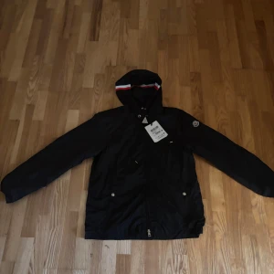 🏆 PRIS KAN DISKUTERAS 🏆 Svart vindjacka från Moncler med huva - Svart vindjacka från Moncler med huva och dragkedja framtill. Jackan har en diskret Moncler-logga på ärmen och en snygg randig detalj i rött, vitt och blått på huvan. Tillverkad i lätt polyester, perfekt för blåsiga dagar.