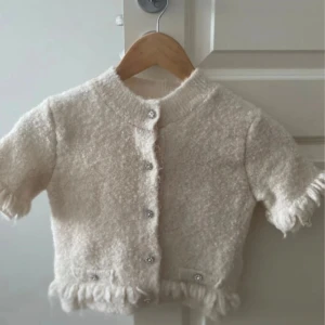 Beige kofta med pärlknappar från H&M - Superfin beige kofta från H&M i kort modell med fluffig känsla. Koftan har korta ärmar, dekorativa pärlknappar och fransdetaljer vid ärmslut och nederkant. Perfekt för dig som gillar en söt och trendig look. Pris går diskutera 