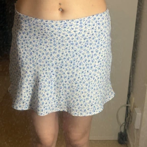 Vit kortkjol med blått blommönster - Supersöt vit kortkjol med små blå blommor och lätt utsvängd form. Perfekt för varma dagar och ger en fräsch och somrig vibe. Kjolen har en mjuk och följsam känsla och passar perfekt till en enkel topp eller stickad tröja.