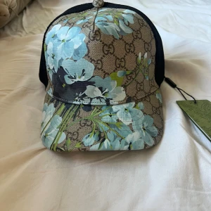 Gucci keps med blå blommor - Snygg keps från Gucci med klassiskt GG-mönster och stora blå blommor framtill. Kepsen har svart mesh baktill och justerbar snapback. Perfekt statement-piece för dig som vill sticka ut. Materialmix med polyester och bomull.