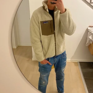 Vit/beige fleecejacka från Patagonia - Säljer en vit/beige fleecejacka från Patagonia med bruna detaljer. Jackan har dragkedja framtill, hög krage och en bröstficka med dragkedja. Perfekt för kyliga dagar och enkel att matcha med jeans eller träningskläder.