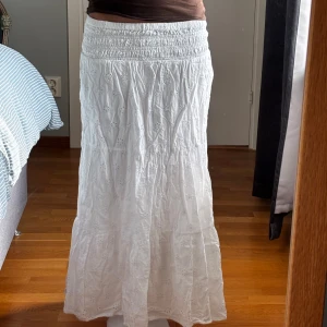 Vit långkjol med broderade blommor - Superfin vit långkjol med broderade blommor och smock i midjan. Kjolen är i bomull och har en luftig och flowy känsla, perfekt för varma dagar. Söt och romantisk stil med små hålbroderier som ger extra detalj.